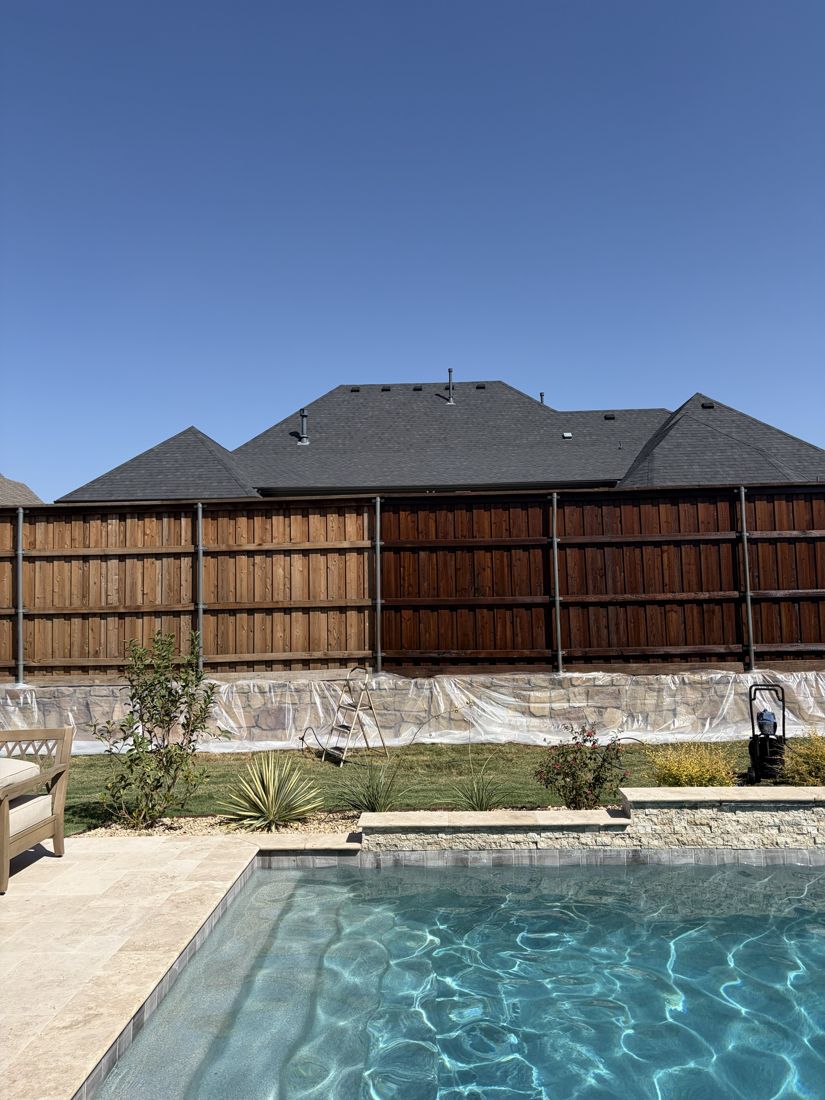 Cedar fence by a pool, left half weathered and unstained, right half freshly stained a deep reddish brown